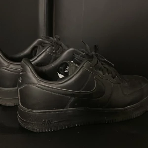 Nike Air Force 1 helsvarta sneakers - Säljer ett par klassiska Nike Air Force 1 i helsvart skinn. Modellen har låg profil, rund tå och snörning. Snygg och stilren design med diskret Nike-logga på sidan och AIR-detalj på hälen. Perfekta för dig som gillar en clean och tidlös look. Pris kan diskuteras!
