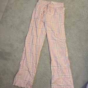 Rosa rutiga byxor från Gina Tricot - Säljer ett par bekväma rosa byxor från Gina Tricot med rutigt mönster. Byxorna har en elastisk midja med snörning för justerbar passform. Perfekta för en avslappnad stil. Omsydda längst ner så de blir längre 