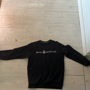Svart Sail Racing sweatshirt - Svart sweatshirt från Sail Racing med vit logga tryckt på bröstet och en mindre logga i nacken. Tröjan har rund halsringning och långa ärmar. Perfekt för dig som gillar en clean och sportig stil.