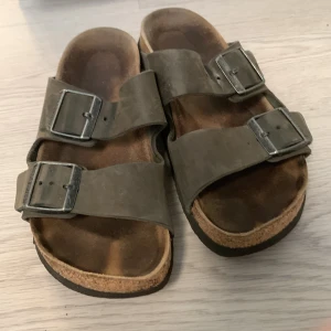  sandaler från Birkenstock - Klassiska sandaler från Birkenstock med två breda remmar i skinn och stora metallspännen. Fotbädden är anatomiskt utformad och sulan är i kork med svart undersida. Perfekta för varma dagar och enkel att matcha med det mesta.