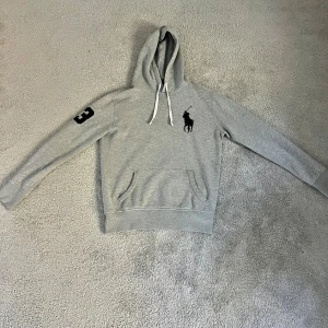 Grå hoodie från Polo Ralph Lauren - Snygg grå hoodie från Polo Ralph Lauren med svart broderad logga på bröstet och nummer 3 på ärmen. Klassisk känguruficka framtill, vita snören i huvan och mjukt material som känns skönt mot huden. Priset är diskuterbart 