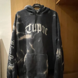 Svart tie-dye Tupac hoodie - Säljer en svart och grå tie-dye hoodie med stor Tupac-print fram och bak samt texten 'All Eyez On Me' på ärmen. Hoodien har huva, känguruficka och långa ärmar. Perfekt för dig som gillar streetwear och hiphop-vibe.
