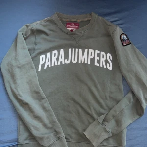 Parajumpers tröja - Snygg grön fleece-tröja från Parajumpers i storlek S. Stor vit logga på bröstet och coolt broderat patch på ärmen. Extra tryck i orange på insidan av nederkanten. Långärmad med ribbade muddar och rund hals. Perfekt för dig som gillar streetwear och vill sticka ut.