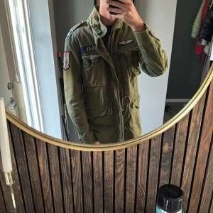 Grön parkajacka Polo Ralph Lauren - Säljer en grön parkajacka från Polo Denim & Supply Ralph Lauren med coola patchar och broderade detaljer på ärmar och bröst. Jackan har flera fickor, snörning i midjan och dragkedja framtill. Perfekt för dig som gillar militärstil och streetwear.