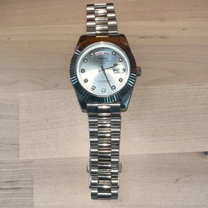 Silverfärgad klocka från Nibosi - Stilren klocka från Nibosi i silverfärg med länkarmband i metall. Urtavlan är också silverfärgad med glittriga timmarkeringar och dag- och datumvisning. Klockan har en klassisk rund form och tydliga detaljer på boetten. Det finns mer länkar till storlek