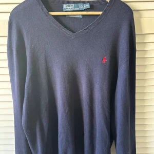 Mörkblå v-ringad tröja Polo Ralph Lauren - Klassisk mörkblå v-ringad tröja från Polo Ralph Lauren i 100% bomull. Tröjan har lång ärm och den ikoniska röda broderade loggan på bröstet. Perfekt för dig som gillar stilrena och tidlösa plagg med premiumkänsla.