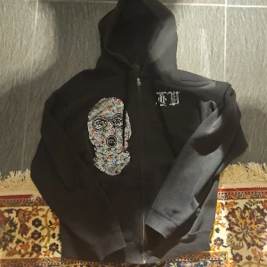 Svart hoodie med strassmask och text - Svart hoodie med dragkedja och huva. På framsidan finns en stor mask i färgglada strasstenar och på ryggen står det 'BANK ROBBER' i strass. Hoodien har långa ärmar och är tillverkad i mjukt material, perfekt för dig som gillar statement-plagg. Det finns lite stenar som har lossnat!!!