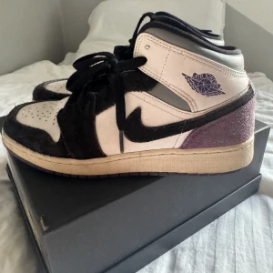 Nike Air Jordan 1 Mid svart/vit/lila - Nike Air Jordan 1 Mid i svart, vit och lila med klassisk Jordan-logga på sidan. Skorna har snörning, svart Swoosh och lila mockadetaljer på hälen samt lila sula. Ovandelen är i skinn och mocka för en snygg kontrast. Perfekta för dig som gillar streetstyle.