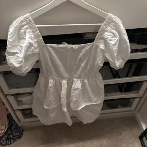 Vit kort klänning med puffärmar - Superfin vit kort klänning med puffiga ärmar och volang i kjolen. Klänningen har fyrkantig urringning och snygg knytning i ryggen. Perfekt för dig som gillar romantisk och trendig stil. Tillverkad i bomull för en luftig känsla.