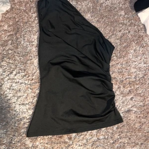 Svart oneshoulder topp - Snygg svart topp med oneshoulder-design och lätt rynkade detaljer på sidan. Perfekt för dig som vill sticka ut med en stilren och trendig look. Toppens material känns mjukt och följsamt mot huden.