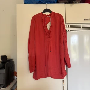 Röd blus med snörning från Odd Molly - Säljer en korallröd långärmad blus/ tunika från Odd Molly med snygg snörning framtill och dekorativa virkade detaljer längs ärmar och rygg. Blusen har en avslappnad passform och är tillverkad i ett  lätt och luftigt tyg, perfekt för varmare dagar.Storlek 1 som enligt deras storleksguide motsvarar en storlek S. Men den är rymlig i storlek. Helt ny med tags/ prislapp fortfarande kvar. Den ör mer en korall/röd ton. Svårt att få rätt bild på färgen. 