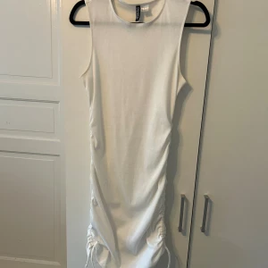 Vit ribbad kortklänning H&M Divided - Snygg vit ribbad kortklänning från H&M Divided. Ärmlös modell med rund hals och cool snörning på båda sidorna som ger en trendig rynkad effekt. Perfekt för dig som gillar enkel men stilren look.
