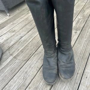 Svarta ridstövlar i skinn från CRW - Snygga svarta ridstövlar från CRW i äkta skinn med dragkedja bak och diskret CRW-logga vid skaftet. Stövlarna har rund tå och klassisk ridstil, perfekta för dig som vill ha en stilren look i stallet.