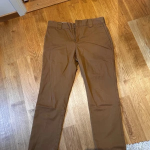 Bruna slim fit chinos från Dickies - Snygga bruna chinos från Dickies i slim fit-modell. Klassisk design med bälteshällor, fickor fram och bak samt diskret Dickies-logga ovanför bakfickan. Tillverkade i slitstark bomull med dragkedja i gylfen. Perfekta för en clean och stilren look.