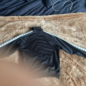 Svart Fred Perry zip-tröja M - Svart zip-tröja från Fred Perry med vit logoband längs ärmarna och broderad Fred Perry-logo på bröstet. Tröjan har hög krage, dragkedja framtill och två fickor. Ingenting fel på tröjan nästan helt nytt skick.