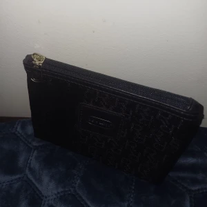 Svart plånbok från Revlon med monogram - Snygg svart plånbok från Revlon med diskret monogrammönster och logga framtill. Väskan har dragkedja upptill. Perfekt att ha med sig i väskan för att hålla ordning på småsaker.