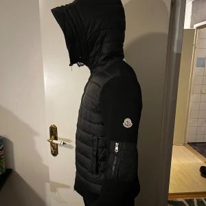 Svart Moncler pufferjacka med huva - Säljer en svart pufferjacka från Moncler med stickade ärmar och vadderad kropp. Jackan har huva, dragkedja framtill och en ficka med dragkedja på ärmen. Klassisk Moncler-logga på armen och snygg, modern look. Perfekt för kalla dagar.
