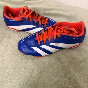 fotbollsskor - Säljer ett par Adidas Predator fotbollsskor i blått med vita detaljer och orange snörning. Skorna har röd insida och yttersula med dobbar för bra grepp på planen. Snygg sportig design med klassiska Adidas-ränder på sidorna. Skorna kommer skickas ut rena.