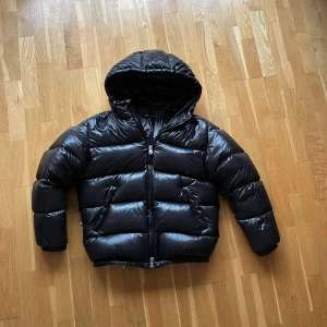 Svart pufferjacka med huva - Säljer en svart pufferjacka från H&M med glansig finish och huva. Jackan har dragkedja framtill, två sidofickor och breda, vadderade sektioner som ger en schysst volym. Perfekt för kalla dagar och riktigt snygg till jeans. Inga hål eller märkbara slitningar. ‼️Pris går såklart att diskutera ‼️