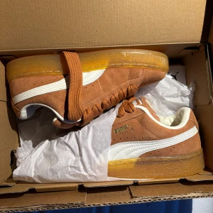 Puma Suede sneakers med gummisula - Snygga Puma Suede sneakers i brun mocka med breda snören och vit sidostrip. Skorna har en genomskinlig gummisula med mönster undertill och gulddetaljer vid loggan. Klassisk rund tå och platt sula. Helt nya och oanvända✨