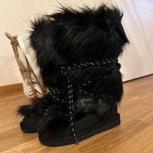 Svarta fluffiga moon boots med snörning - Säljer ett par svarta moon boots med lång, fluffig pälsimitation och coola snören runt skaftet från Nakd. Skorna har platt sula och är perfekta för vintern när du vill sticka ut. Riktigt mysiga och unika boots som håller dig varm och snygg. 