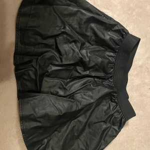 Svart kortkjol i skinnimitation från Lindex - Svart kortkjol från Lindex med bred resår i midjan. Kjolen har en cool skinnlook och är lätt utställd för en snygg siluett. Perfekt för dig som gillar en edgy stil och vill sticka ut lite extra.