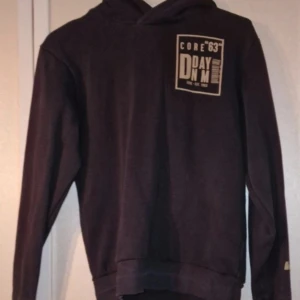 Svart hoodie med tryck - Svart hoodie från DMD med tryck framtill och stort tryck med streckkod på ryggen. Klassisk huva, ribbade muddar och mjukt material. Perfekt för dig som gillar streetstyle och vill sticka ut med detaljer. Den kan möjligtvis ha blivit något blekt i färgen efter tvätt och den har fått en rand på mudden (se bild). Inga andra skavanker vad jag har kunnat se. Stl 146/152