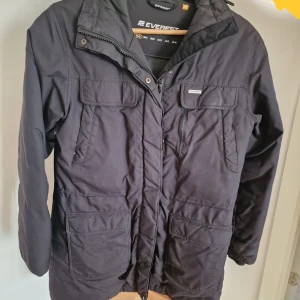 Svart parkajacka från Everest med fuskpäls - Svart parkajacka från Everest med huva och fuskpäls, flera praktiska fickor framtill och dragkedja med knappslå. Jackan är långärmad och har en klassisk parkamodell som håller dig varm under kalla dagar.