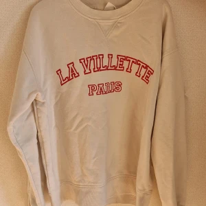Vit sweatshirt La Villette Paris - Säljer en vit sweatshirt med röd text 'La Villette Paris' framtill. Tröjan har rund halsringning, ribbade muddar och en avslappnad passform. Perfekt för dig som gillar en enkel men snygg stil med fransk vibe.