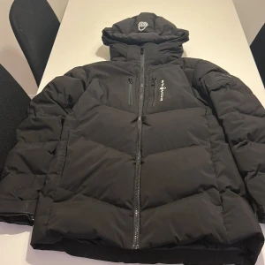 Svart Sail Racing jacka med luva  - Säljer en svart Sail Racing jacka från Sail Racing med dragkedja fickor. Jackan har ett stilrent, quiltatmönster och logga på armen. Jättebra för kalla dagar och väldigt snygg.