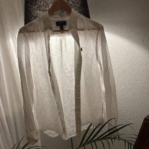 Vit linneskjorta från Polo Ralph Lauren - Vit skjorta i tunn och luftig linne från Polo Ralph Lauren. Klassisk krage, långärmad och med knappar framtill. Diskret broderad logga i marinblått på bröstet. Perfekt för en chill och clean look under varmare dagar.