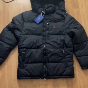 Svart pufferjacka från Polo Ralph Lauren - Svart pufferjacka från Polo Ralph Lauren med huva och dragkedja framtill. Jackan har två sidofickor med dragkedja och en röd broderad logga på bröstet. Materialet är en blandning av bomull och polyamid, vilket gör den både varm och lätt. Perfekt för kalla dagar.