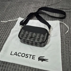 Lacoste väska - Snygg axelväska från Lacoste med ett geometriskt mönster i svart och grått. Väskan har en justerbar axelrem och en dragkedja för säker förvaring. Perfekt för att bära dina nödvändigheter med stil. Den går för 1600kr 