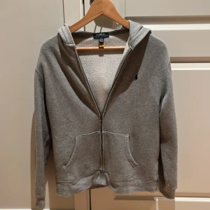 Grå hoodie från Polo Ralph Lauren XL - Grå hoodie från Polo Ralph Lauren i storlek XL barn / xs/ S i vuxen med dragkedja och huva. Tröjan har två stora fickor framtill och ett litet broderat logomärke på bröstet. Insidan är mjuk och fleecefodrad, perfekt för kyliga dagar. Klassisk och enkel design som funkar till allt.