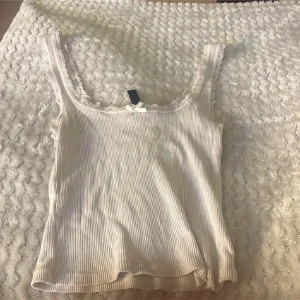 Vit ribbad linnetopp med spets - Supersöt vit linnetopp med ribbad struktur och spetskant runt halsringningen. Dekorerad med en liten rosett framtill för extra gullig vibe. Toppen är ärmlös och har en mjuk, lätt stretchig känsla som gör den skön att bära. Perfekt för sommarens alla dagar!