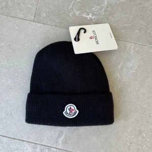 Svart Moncler mössa med fleecefoder - Svart ribbstickad mössa från Moncler med klassisk logotyp framtill. Insidan är fodrad med mjuk vit fleece för extra värme och komfort. Perfekt för kalla dagar och har en enkel, stilren design som funkar till alla jackor.