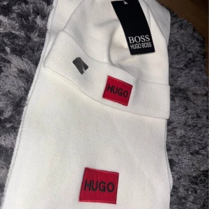 Vit halsduk från Hugo Boss - Stilren vit halsduk från Hugo Boss med röd HUGO-logga broderad på ena änden. Halsduken är mjuk och har en klassisk, avlång form som passar perfekt till kalla dagar. Snygg accessoar för att lyfta din vinterstil.