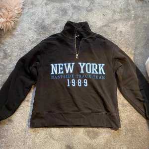 Svart half zip sweatshirt New York - Svart sweatshirt med half zip och hög krage. Tryck i blått och vitt framtill med texten 'NEW YORK EASTSIDE TRACK TEAM 1989'. Mjuk och skön, perfekt för chill dagar. Långärmad och ribbade muddar vid ärmslut och nederkant.