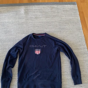 Mörkblå sweatshirt från GANT - Säljer en mörkblå sweatshirt från GANT med broderad logga och sköld på bröstet. Tröjan har rund halsringning, långa ärmar och ribbade muddar. Perfekt för en stilren och sportig look.