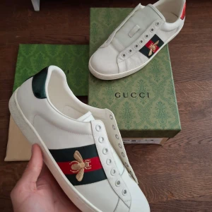 Gucci Ace sneakers med bi broderi - Gucci Ace sneakers i vitt skinn med klassiska gröna och röda ränder på sidan, dekorerade med en broderad guldfärgad bi. Baksidan har metallicröd och metallicgrön detalj med Gucci-logga. Platt sula och rund tå. Allt og medföljer. Skick 9/10. Priset är inte hugget I sten.