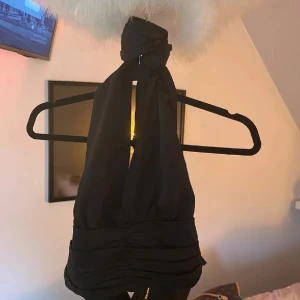 Svart festtopp med öppen rygg - Säljer en svart halterneck topp med breda axelband som knyts i nacken. Toppen har en djup v-ringning och snygga rynkade detaljer framtill. Perfekt för dig som vill ha en stilren och trendig look.