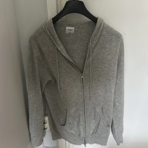 Grå kashmirhoodie från John Henric - Mjuk och stilren grå hoodie från John Henric i lyxig kashmir. Tröjan har dragkedja framtill, två fickor, snörning i huvan och långa ärmar. Perfekt för dig som gillar en clean och chill look. Tröjan har dock ett hål i ärmen vilket är varför det billiga priset är.