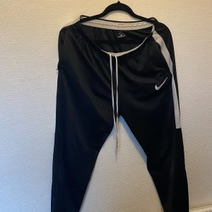 Nike Dri-Fit svarta träningsbyxor - Nike Dri-Fit träningsbyxor i svart med vita detaljer längs sidorna och vit snörning i midjan. Byxorna har elastisk midja och är gjorda i ett lätt syntetmaterial som andas. Swoosh-logga på benet för sportig vibe.