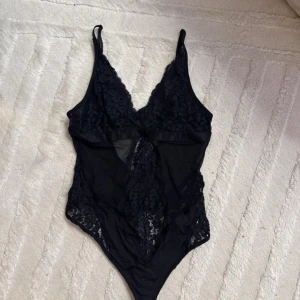 Svart spetsig korsett från Hunkemöller - Svart korsett från Hunkemöller i storlek XS med tunna axelband och v-ringad spets upptill. Snygga spetsdetaljer och meshpaneler framtill ger en extra cool vibe. Perfekt för dig som gillar att sticka ut med detaljerad design och feminin touch.