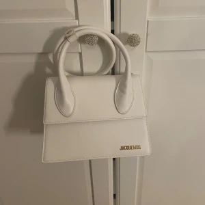 Vit handväska Jacquemus  - Säljer en stilren vit handväska från Jacquemus med guldiga detaljer och logga framtill. Väskan har både handtag och justerbar axelremm.Den har en snygg, srilren och är köpt för 1000kr knappt använd max 2 gånger inga tecken på användning