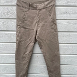 Beige byxor från Kappahl, strl 38 - Snygga beige byxor från Kappahl med hög midja och raka smala ben i kortare modell. . Byxorna har fickor fram och fusk bak, bälteshällor och stängs med dragkedja, knapp och hyska. Materialet är mjukt med 76% viskos, 21% polyamid och 3% elastan för skön stretch. Använda en gång. 