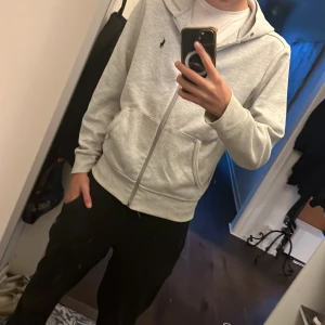 Grå hoodie från Polo Ralph Lauren - Snygg ljusgrå hoodie från Polo Ralph Lauren med klassisk svart logga på bröstet. Tröjan har huva, dragkedja framtill och två fickor. Perfekt för chill dagar och enkel att matcha med jeans eller joggers.