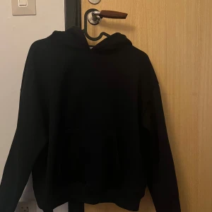 Svart hoodie med huva - En klassisk svart hoodie med huva och magficka. Tröjan har en avslappnad passform och långa ärmar, perfekt för att chilla eller dra på sig över en t-shirt. Enkel och stilren design utan tryck eller detaljer.