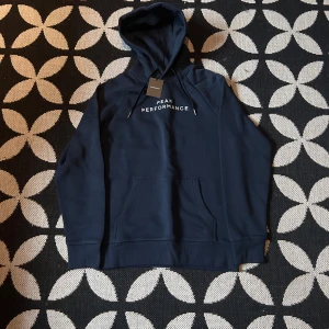 Mörkblå hoodie Peak Performance S - Helt ny aldrig använt. Mörkblå hoodie från Peak Performance i storlek S. Tröjan har huva med snörning, broderad logga på bröstet och en stor magficka. Tillverkad i mjuk bomullsmix, perfekt för chill eller träning. Klassisk och stilren design med normal passform.
