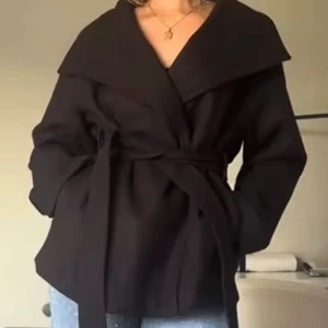 Svart omlottkappa med bälte - Stilren svart kappa med bred krage och knytskärp i midjan. Jackan har omlottmodell och långa ärmar, vilket ger en snygg och avslappnad siluett. Perfekt för dig som gillar minimalistisk och trendig look.